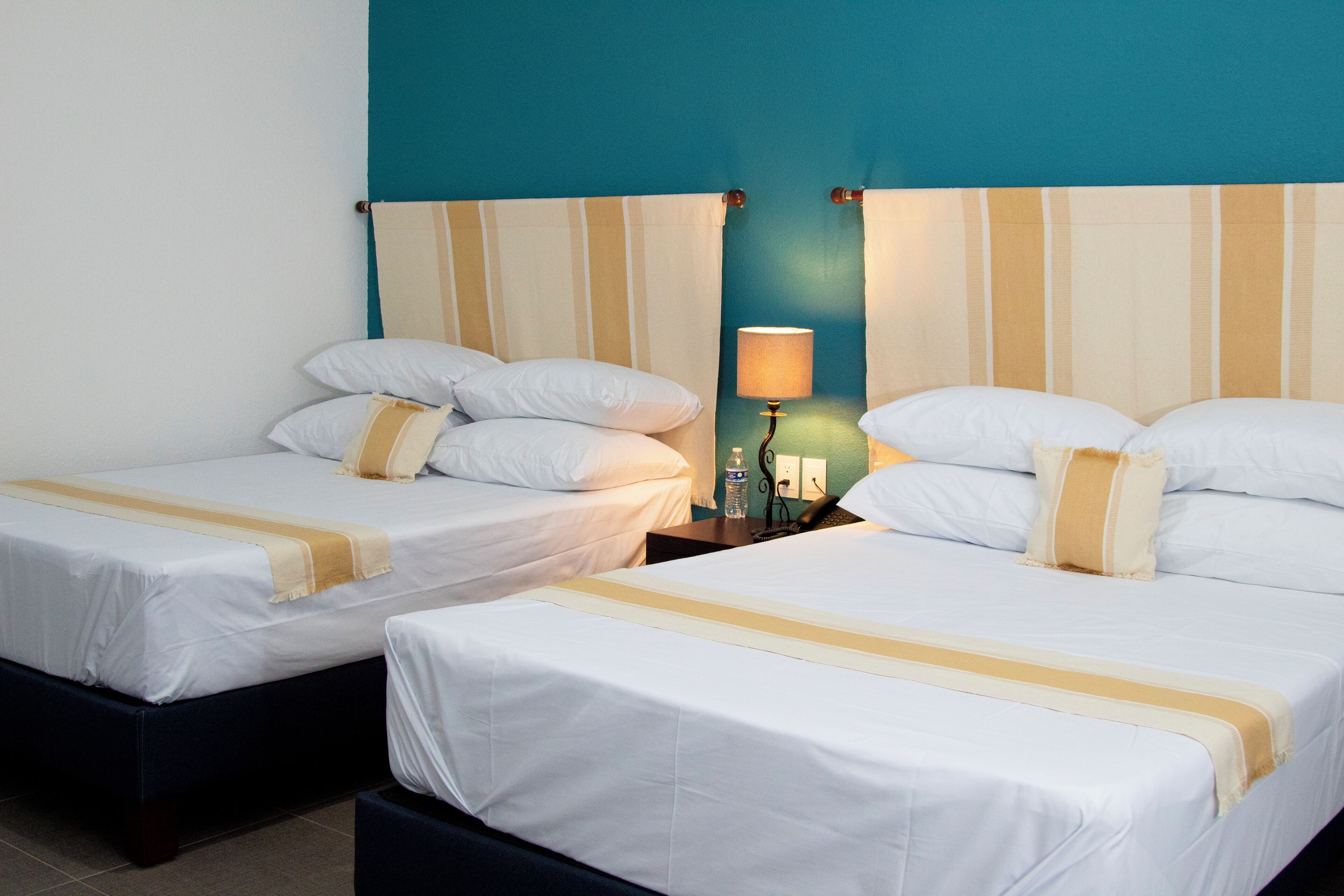 Deux lits simples avec du linge de lit blanc et des touches de couleur beige, une table de chevet avec une lampe, et un mur de couleur turquoise.