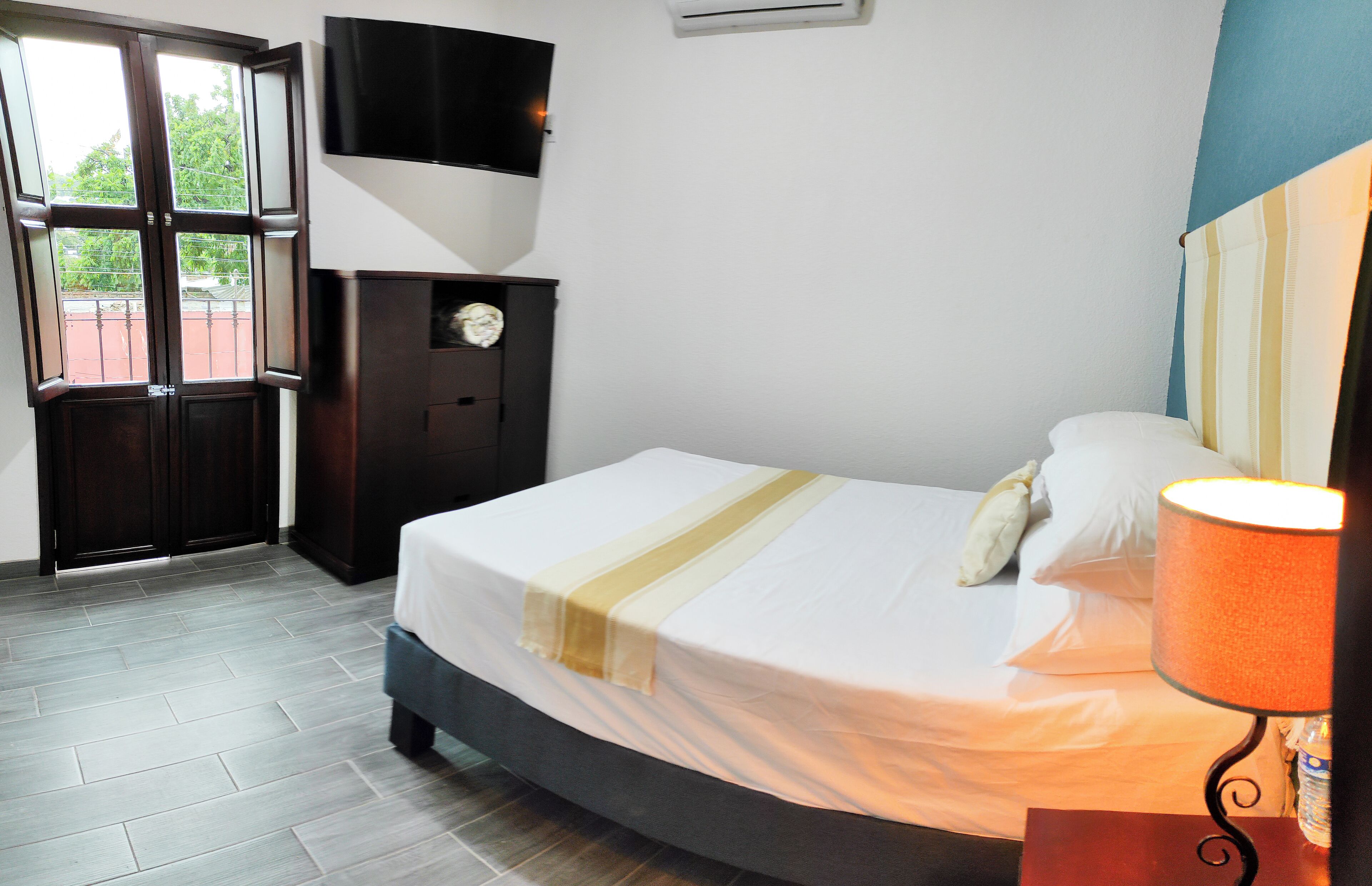Habitación de hotel con una cama, un televisor montado en la pared, una mesita de noche con lámpara y una puerta que da a un espacio exterior.