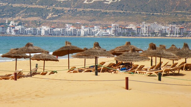 Una playa con sombrillas de paja y tumbonas, una orilla arenosa y una colina al fondo con escritura árabe.