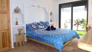Un dormitorio con una colcha azul estampada, un cabecero de madera, un pequeño taburete de madera y vistas a un jardín a través de una puerta corrediza.