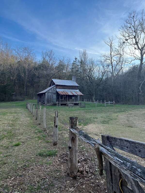 A rustic wooden barn with a metal roof, surrounded by a fenced field and trees.