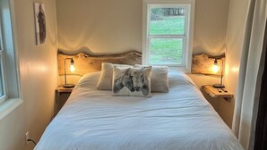 Une chambre dotée d’un grand lit, de deux lampes de chevet, d’une fenêtre donnant sur un paysage verdoyant et d’une œuvre d’art accrochée au mur.