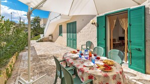 Un área de comedor al aire libre con una mesa preparada para dos, cubierta por un paraguas blanco, contra un fondo de paredes de piedra y contraventanas verdes.