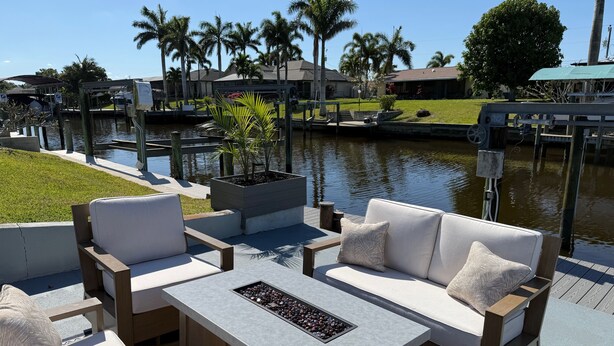 An outdoor seating area with a table and chairs, a planter with a small tree, and a view of a water body and buildings in the background.