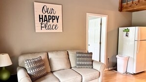 Una sala de estar con sofá color beige, cojines estampados, un letrero en la pared que dice "Our Happy Place", una puerta y un refrigerador blanco.