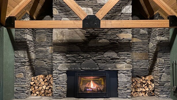 Una chimenea de piedra con fuego, flanqueada por leña apilada, en una habitación con pared de piedra y vigas de madera.