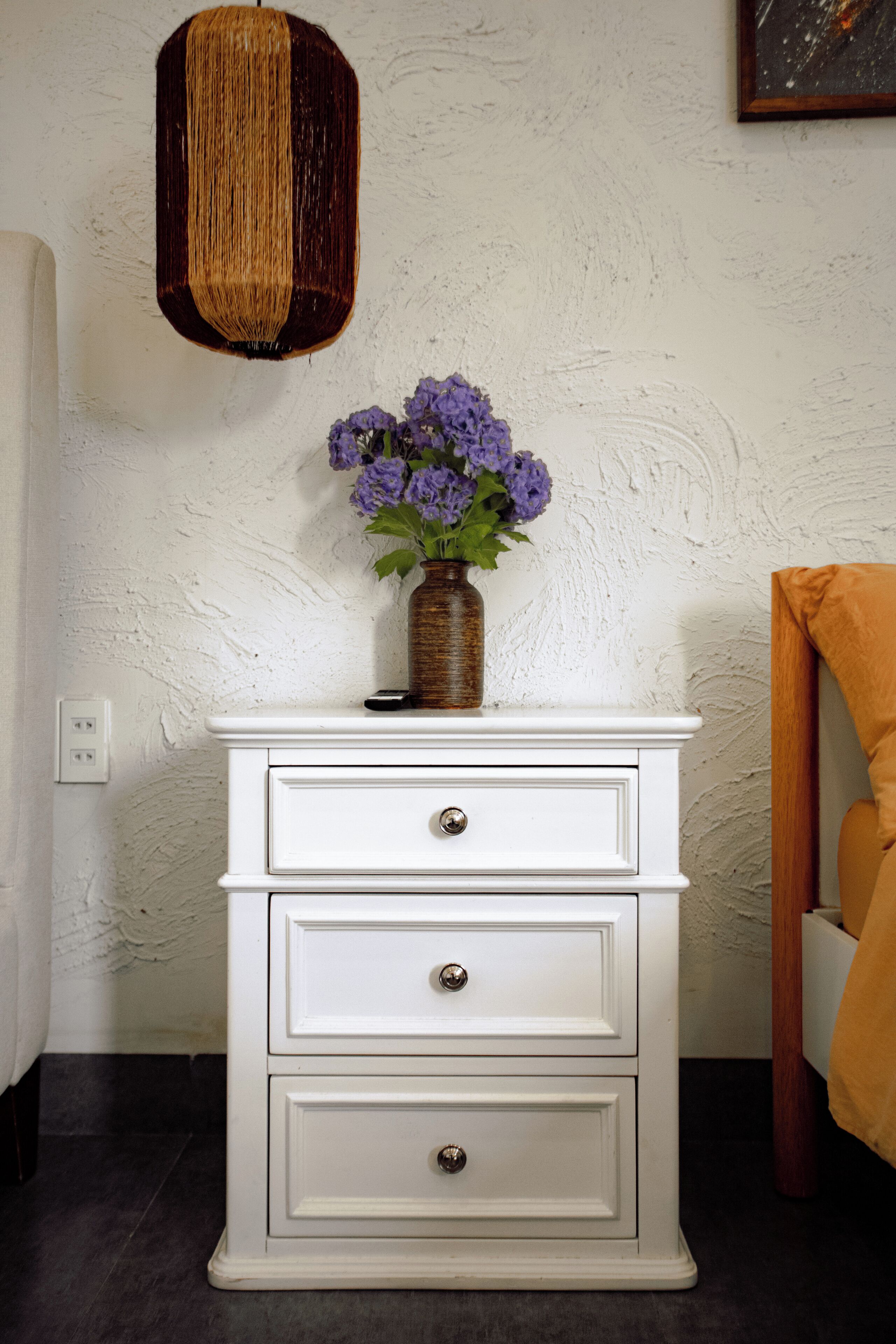 A white dresser with three drawers, a vase with purple flowers, and a textured wall.