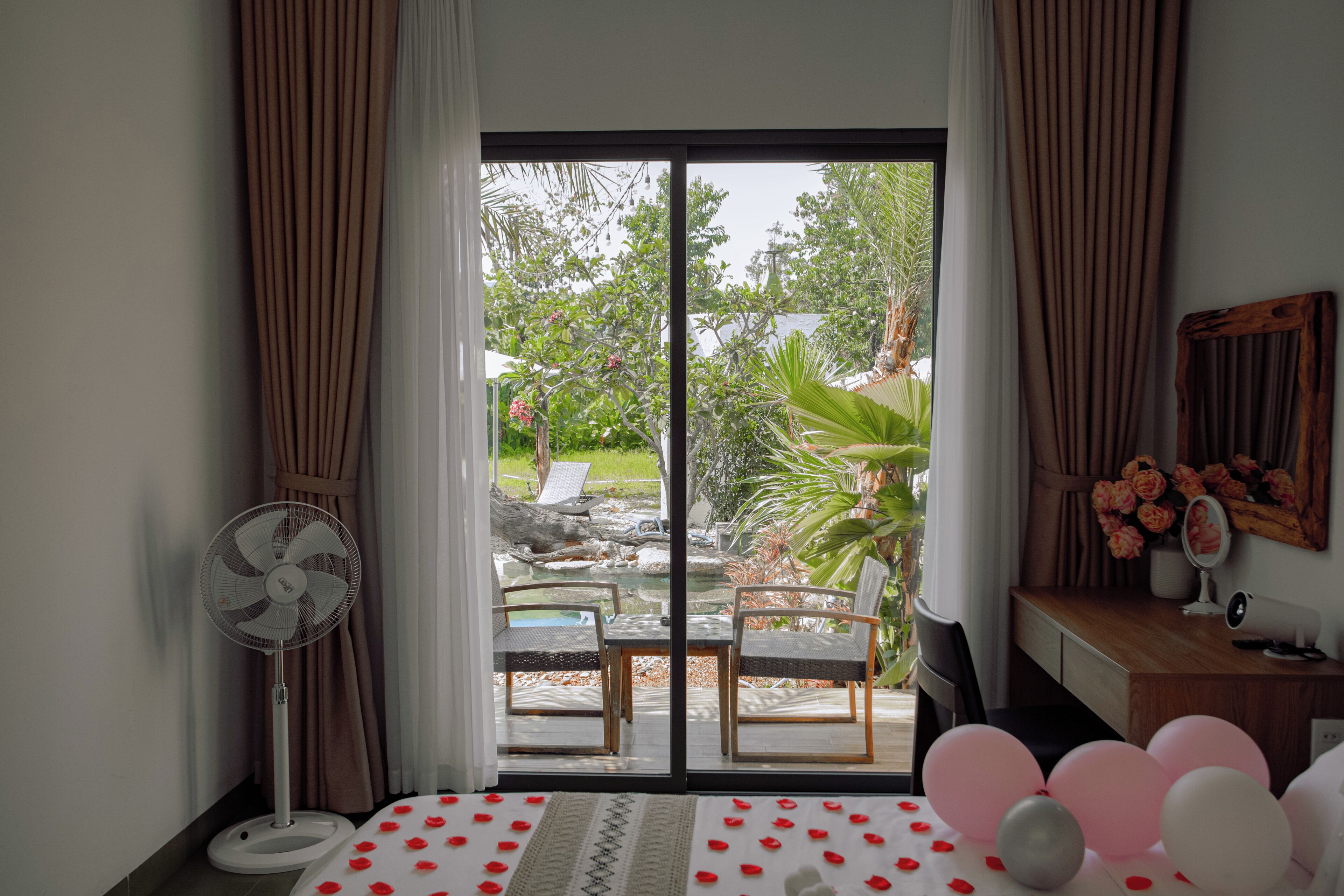 A room with a bed, a fan, a desk, a mirror, and a view of an outdoor patio with chairs and plants.