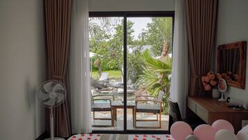 A room with a bed, a fan, a desk, a mirror, and a view of an outdoor patio with chairs and plants.