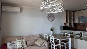 A living room with a sofa, a dining table with chairs, a hanging light fixture, a clock on the wall, and a kitchen area in the background. - Beachfront Four bedroom Apartment (Sveti Stefan)