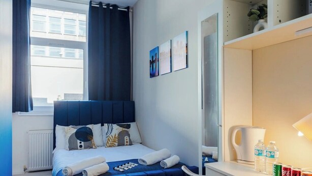 Una camera d'albergo con un letto, una scrivania con lampada, uno specchio e uno scaffale con bottiglie d'acqua e lattine.