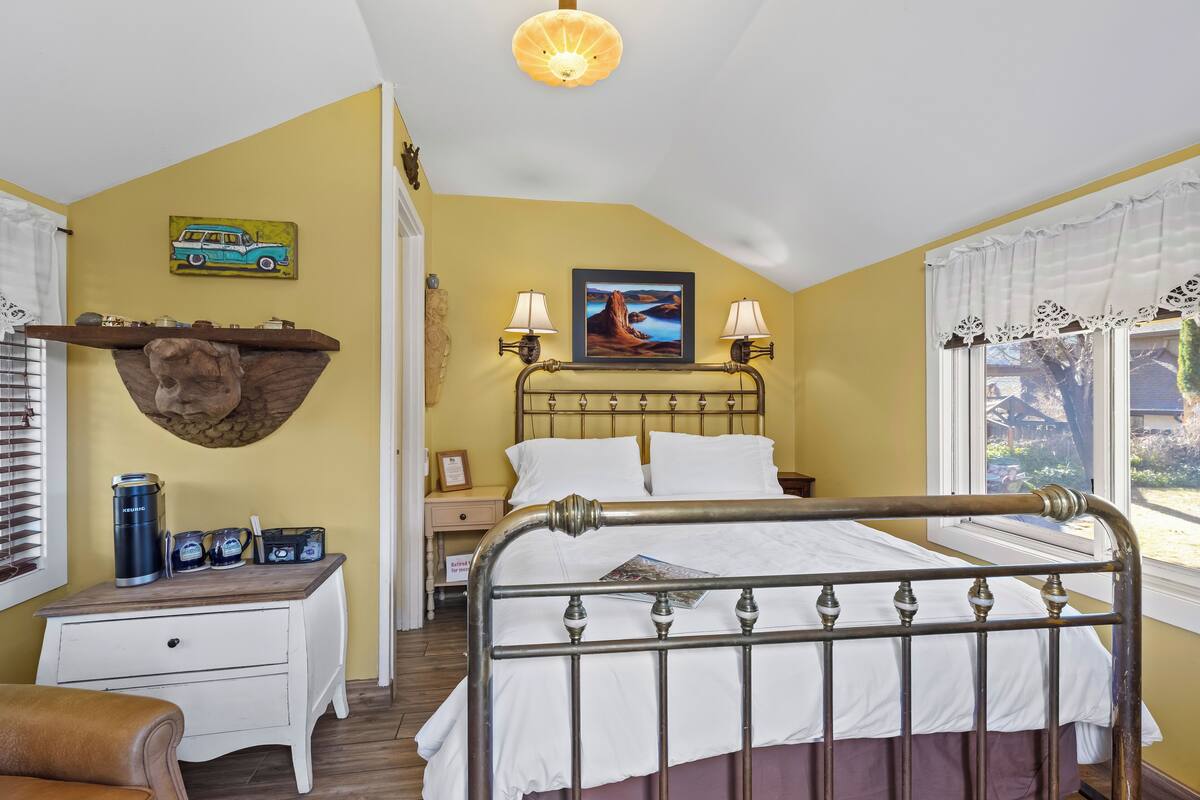 Een slaapkamer met een bed, nachtkastjes, een lamp, een wandplank met een beerbeeld en een raam met gordijnen.