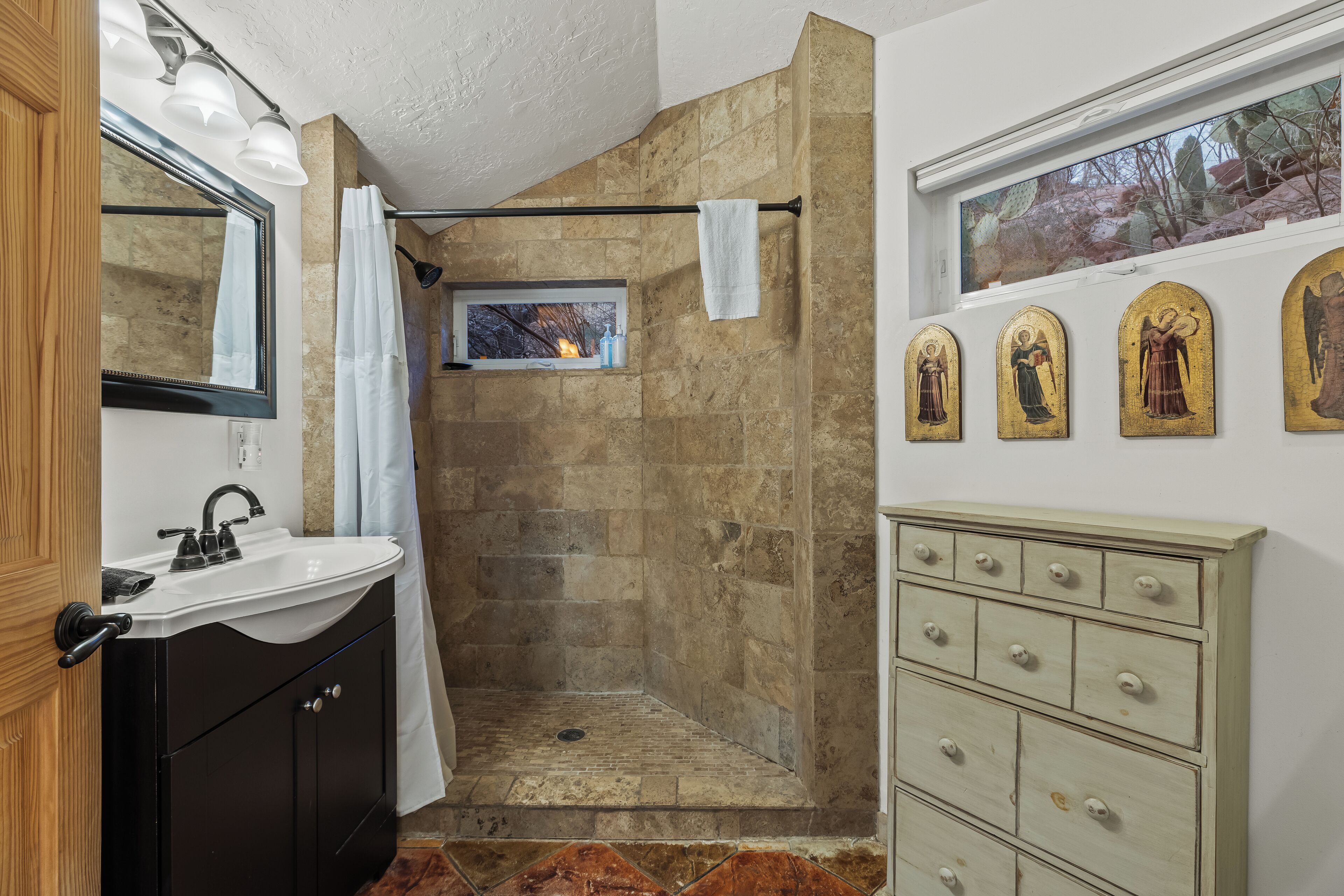 A bathroom with a stone shower, a sink with a mirror, and a vanity with drawers.
