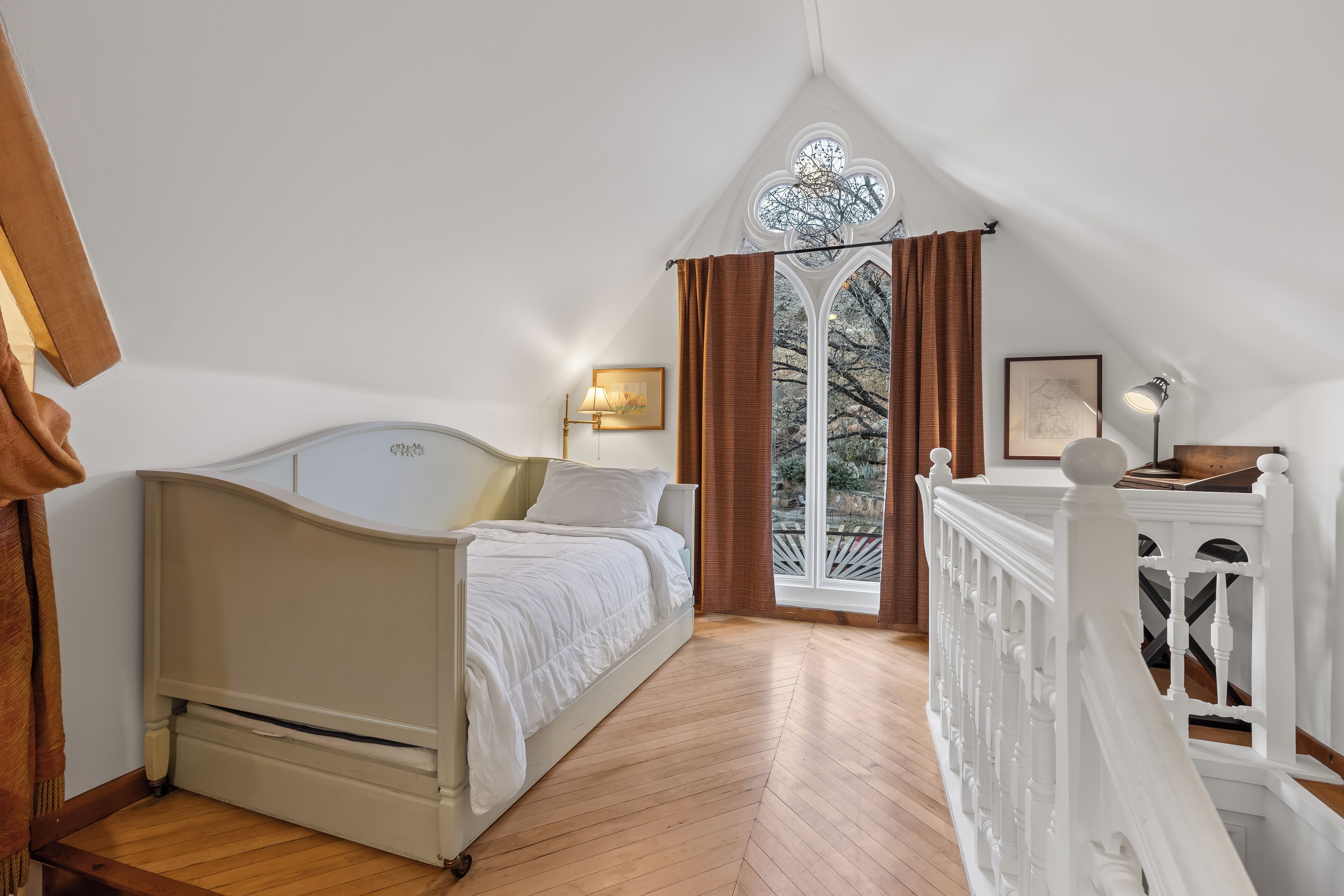 A bedroom with a bed, a window with curtains, a wooden floor, and a white railing.