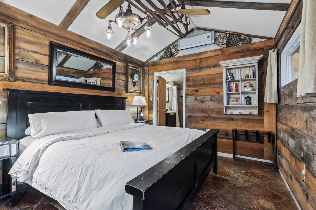 Een slaapkamer met houten inrichting, een bed met witte beddengoed, een nachtkastje, een spiegel, een plafondventilator en een boekenkast.
