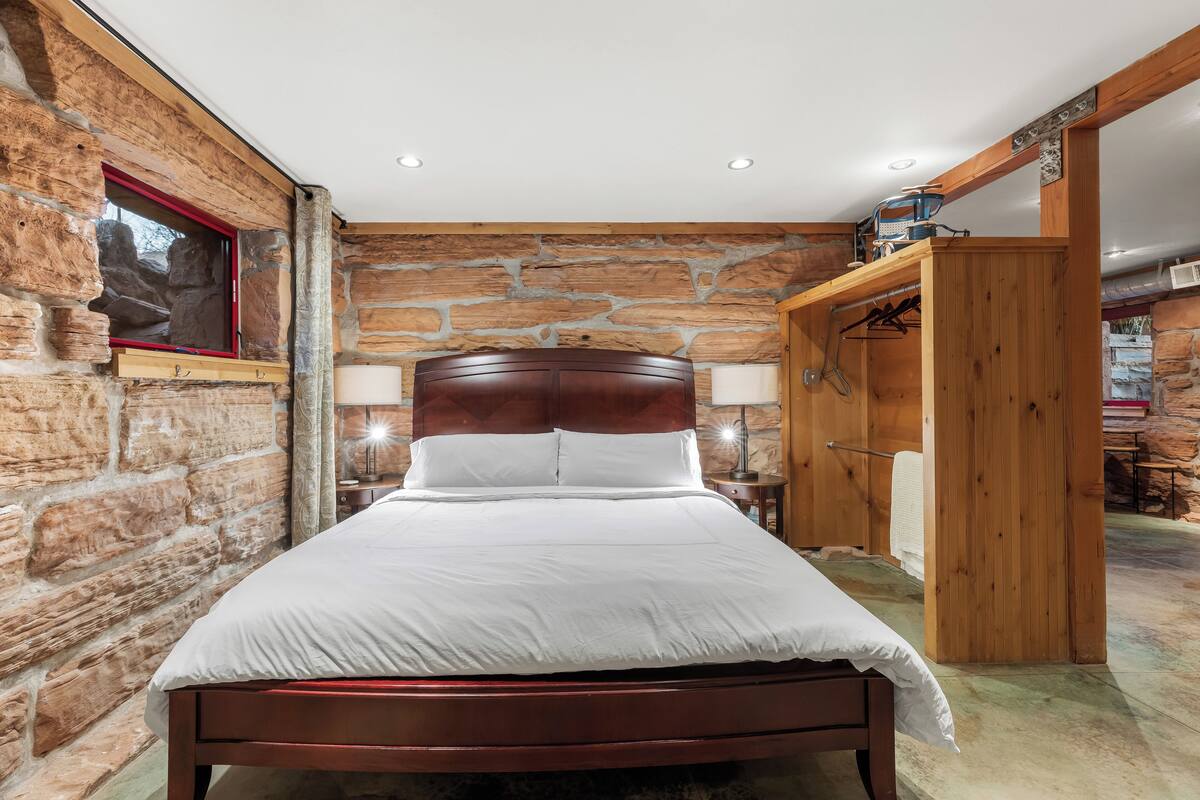 Een slaapkamer met een stenen muur, een houten bed, een houten kledingkast en een raam met rode kader.