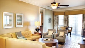 A hotel room with a sofa, armchairs, a coffee table, a ceiling fan, and a dining area with chairs and a table.