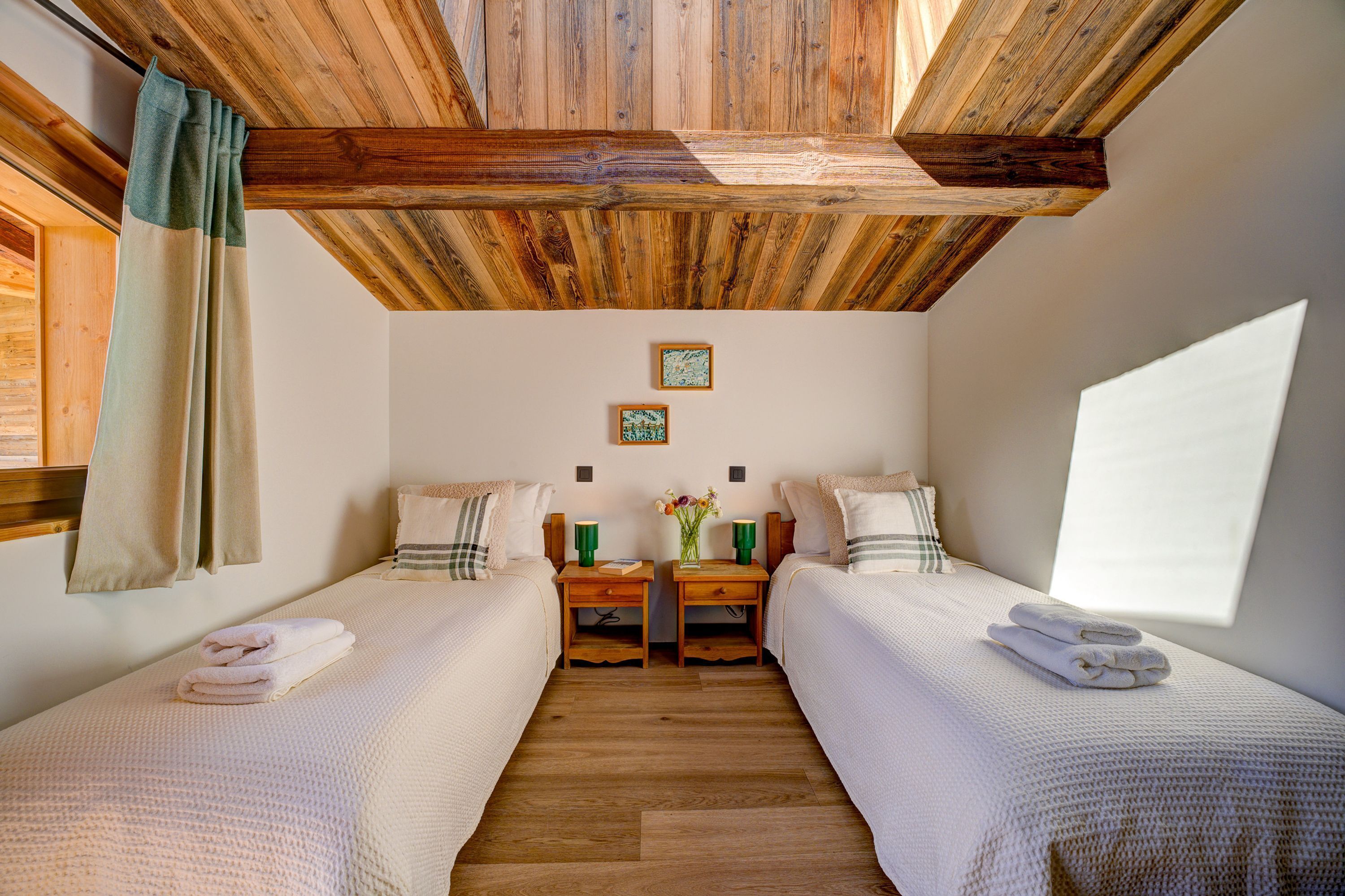 A room with two beds, wooden beams on the ceiling, a small table with a vase, and a window providing natural light.