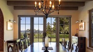 Ein Esszimmer mit langem Tisch, Stühlen, einer Chandelüstere und großen Fenstern mit Blick auf Grünflächen.