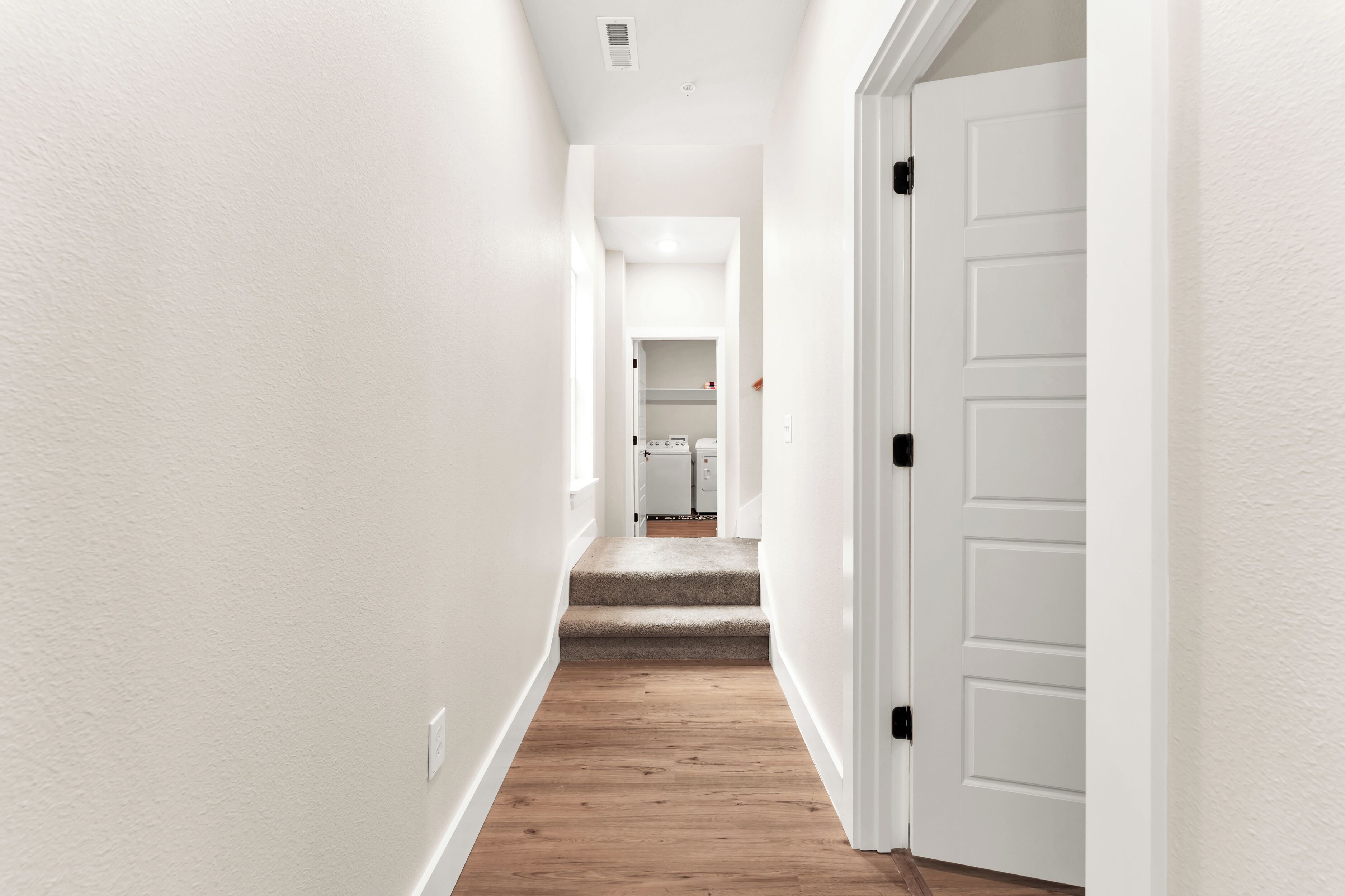 A narrow hallway with white walls and wooden flooring, leading to a door on the right.