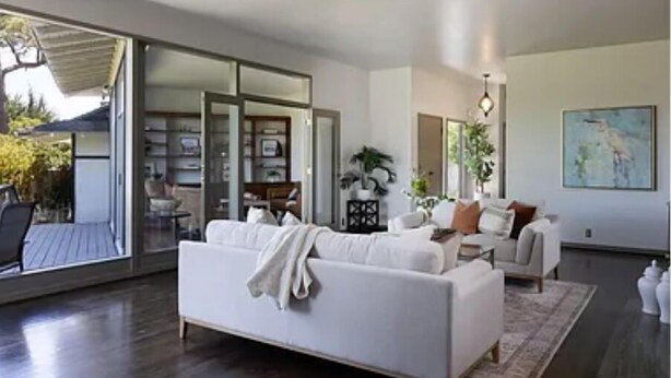 A modern living room with a white sofa, a coffee table, and a dining area with chairs. There is a sliding glass door leading to an outdoor space.