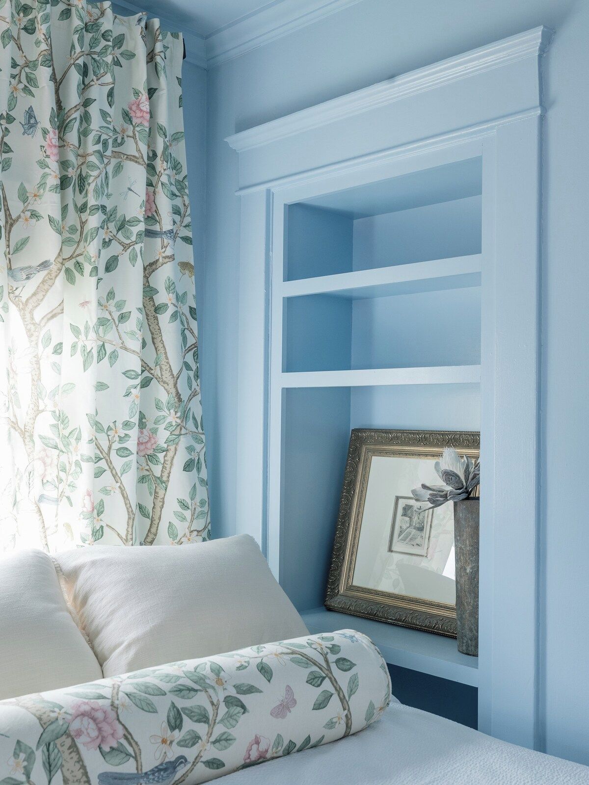 Ein Zimmer mit blauer Wand, einem weißen Sofa mit Blumenkissen und einem gerahmten Bild auf einem Regal.