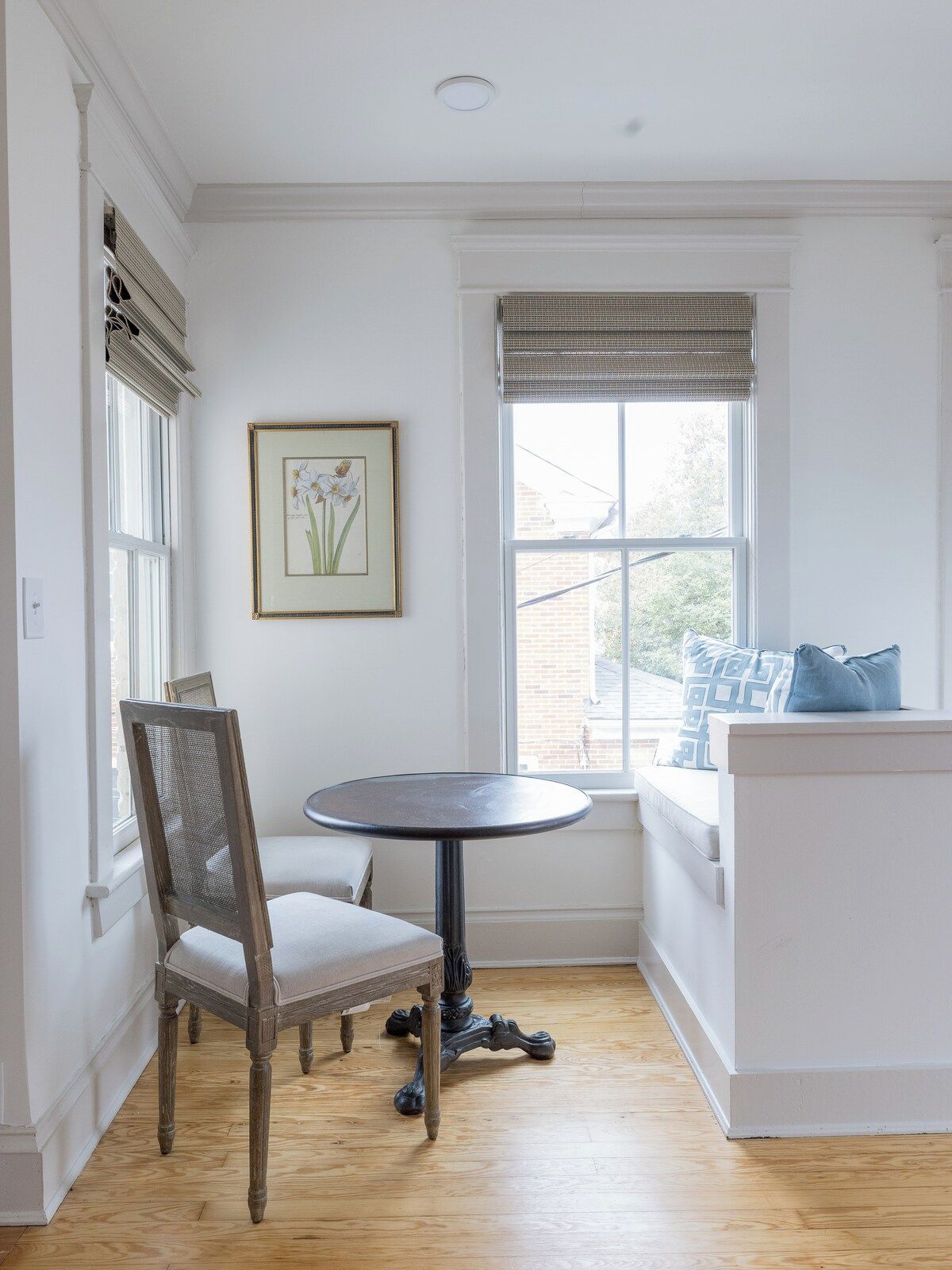 Een kamer met een ronde tafel, een stoel, een raam met jaloezieën en een ingelijst botanisch reproductie aan de muur.