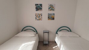 Ein Zimmer mit zwei Einzelbetten, jeweils mit einem türkisfarbenen Kopfteil, weißer Bettwäsche und einem kleinen Tisch dazwischen. An der Wand oberhalb der Betten hängen vier gerahmte Kunstwerke.