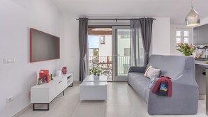 A modern living room with a grey sofa, a white coffee table, and a TV mounted on the wall. A balcony with a table and chairs is visible through the sliding glass door.