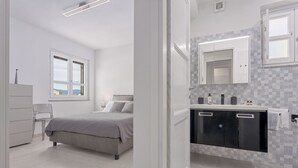 A modern bedroom with a bed, bedside table, and a chair. It features a large window and a bathroom with a mirror, sink, and cabinets.