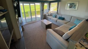 A spacious living area with a large sofa, a coffee table, and a television. There is a sliding glass door leading to a balcony with a view of a grassy field. - The Lodge at Pennant-igyn (Pwllheli)