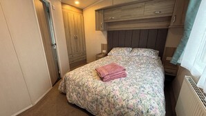 A bedroom with a bed, a nightstand, a wooden wardrobe, and a window with curtains. - The Lodge at Pennant-igyn (Pwllheli)