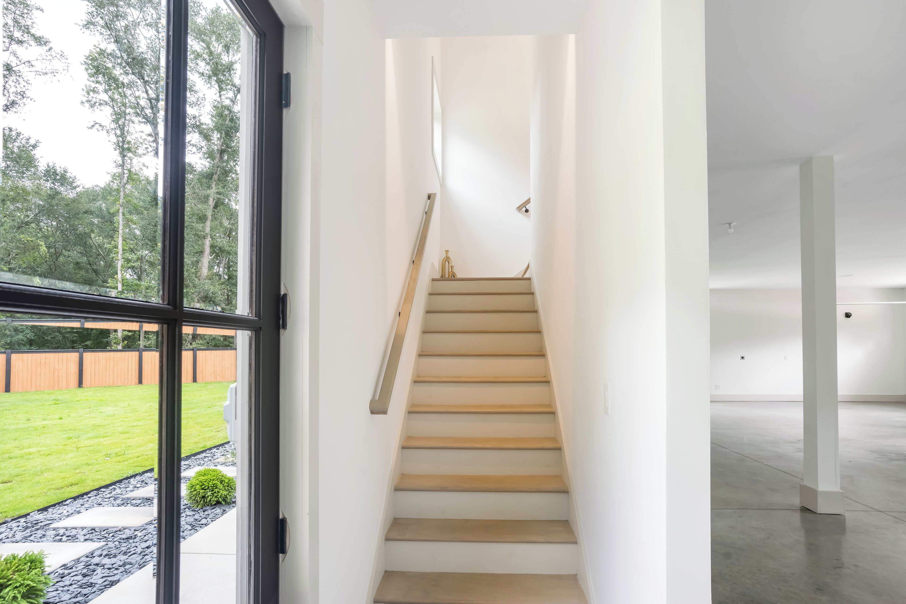 Un interior modern amb una escala que condueix a un segon pis, una finestra gran amb vista a un prat verd, i una habitació espaiosa i buida.