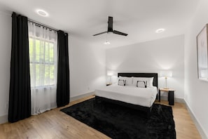 Une chambre à coucher moderne avec un grand lit, deux tables de chevet, un ventilateur de plafond et une fenêtre avec des rideaux légers.