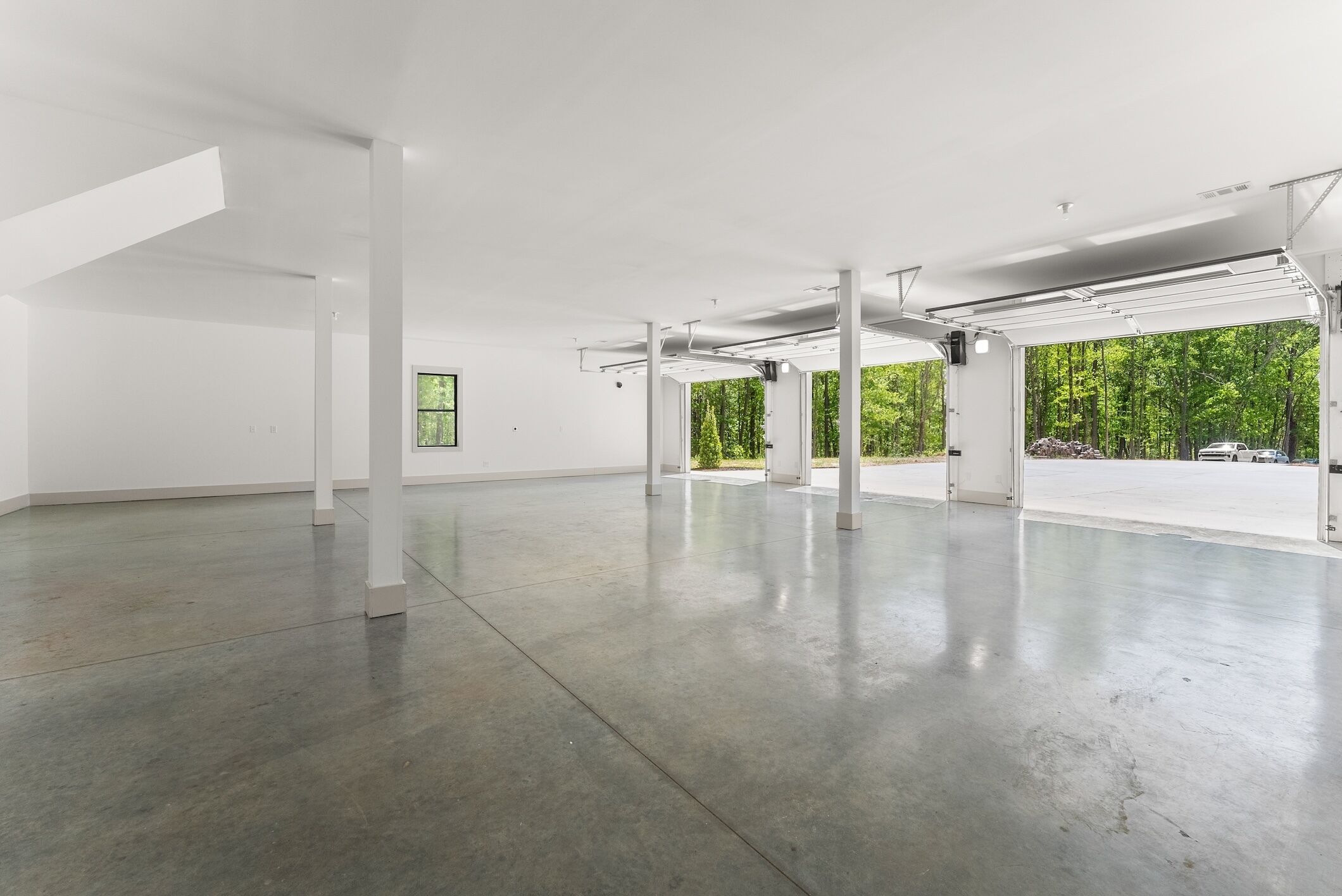 A spacious, empty room with white walls and a concrete floor, featuring large windows and a view of greenery outside.
