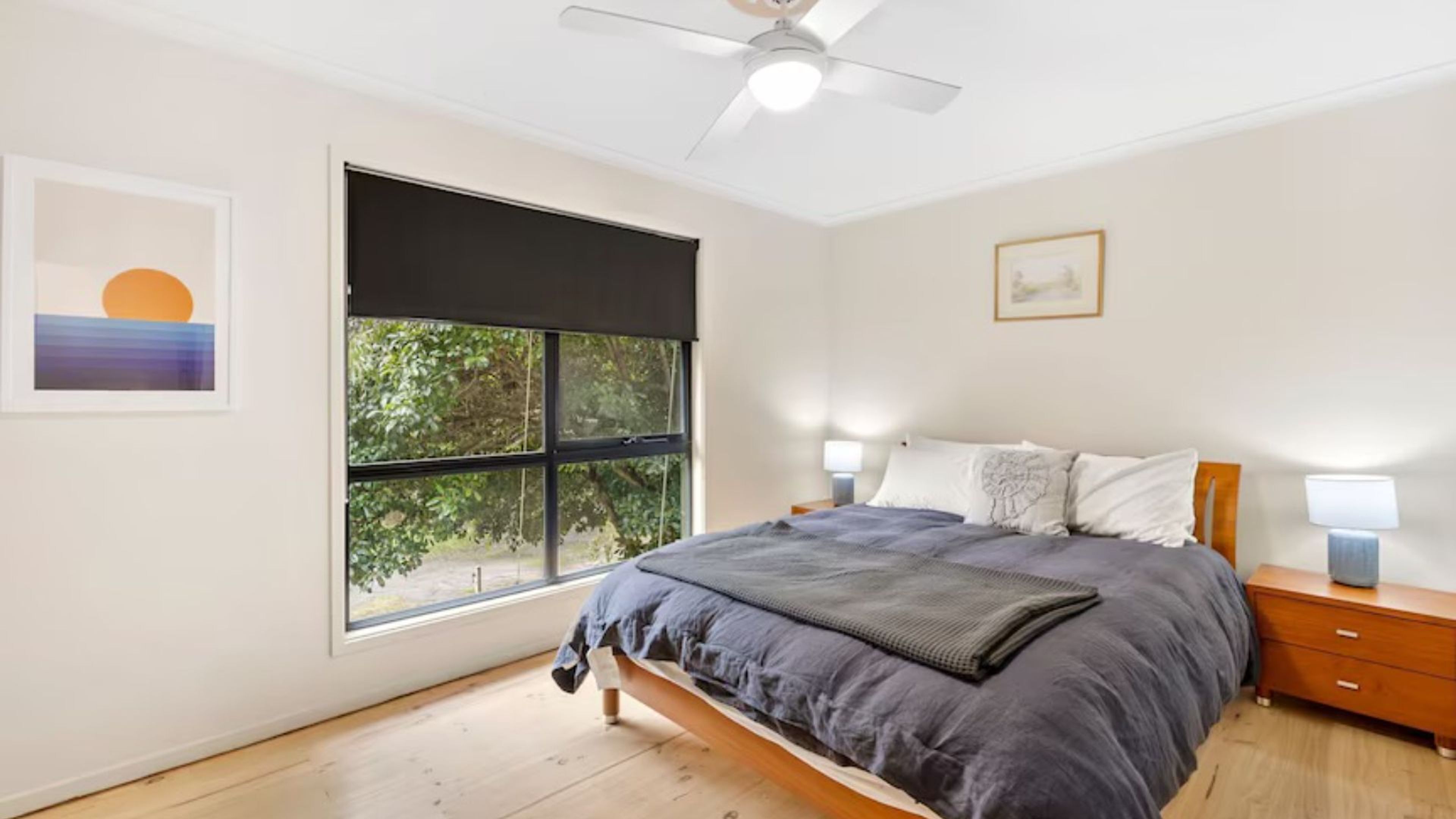 A bedroom with a bed, bedside lamps, a ceiling fan, a window with a view, and artwork on the walls.