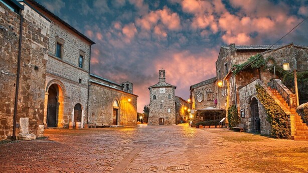 Una calle adoquinada flanqueada por edificios históricos de piedra, una torre del reloj central e iluminación ambiental cálida.
