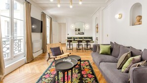 A modern living room with a grey sofa, a wooden chair, a dining table with chairs, and a colorful rug. - A PERFECT PARISIAN GETAWAY IN A PERFECT LOCATION – RUE DU TEMPLE (Paris)