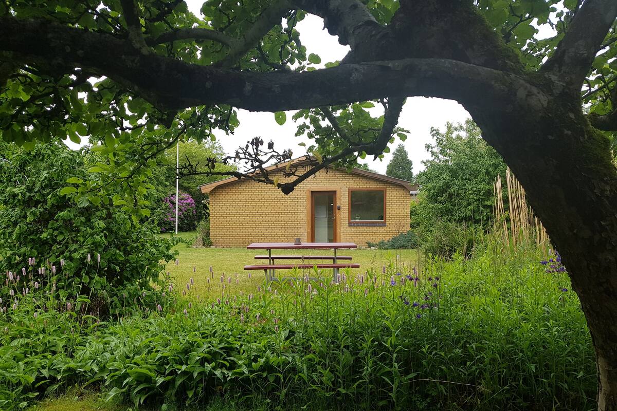 A small brick building with a door and windows, surrounded by lush greenery and a picnic table in the foreground.