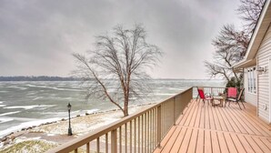 A wooden deck with a railing, overlooking a frozen body of water with trees and a house in the background.