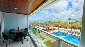A balcony with a view of a swimming pool, outdoor seating, and palm trees. - Solar Tambaú Luxo - By Almare Flats (João Pessoa)