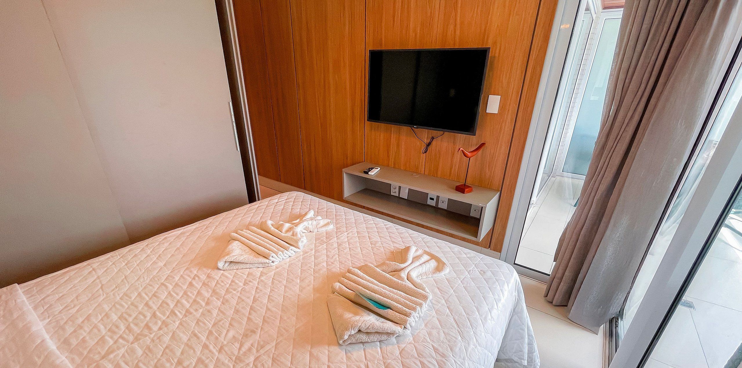 A hotel room with a bed, a TV mounted on the wall, and a small shelf with a remote control.