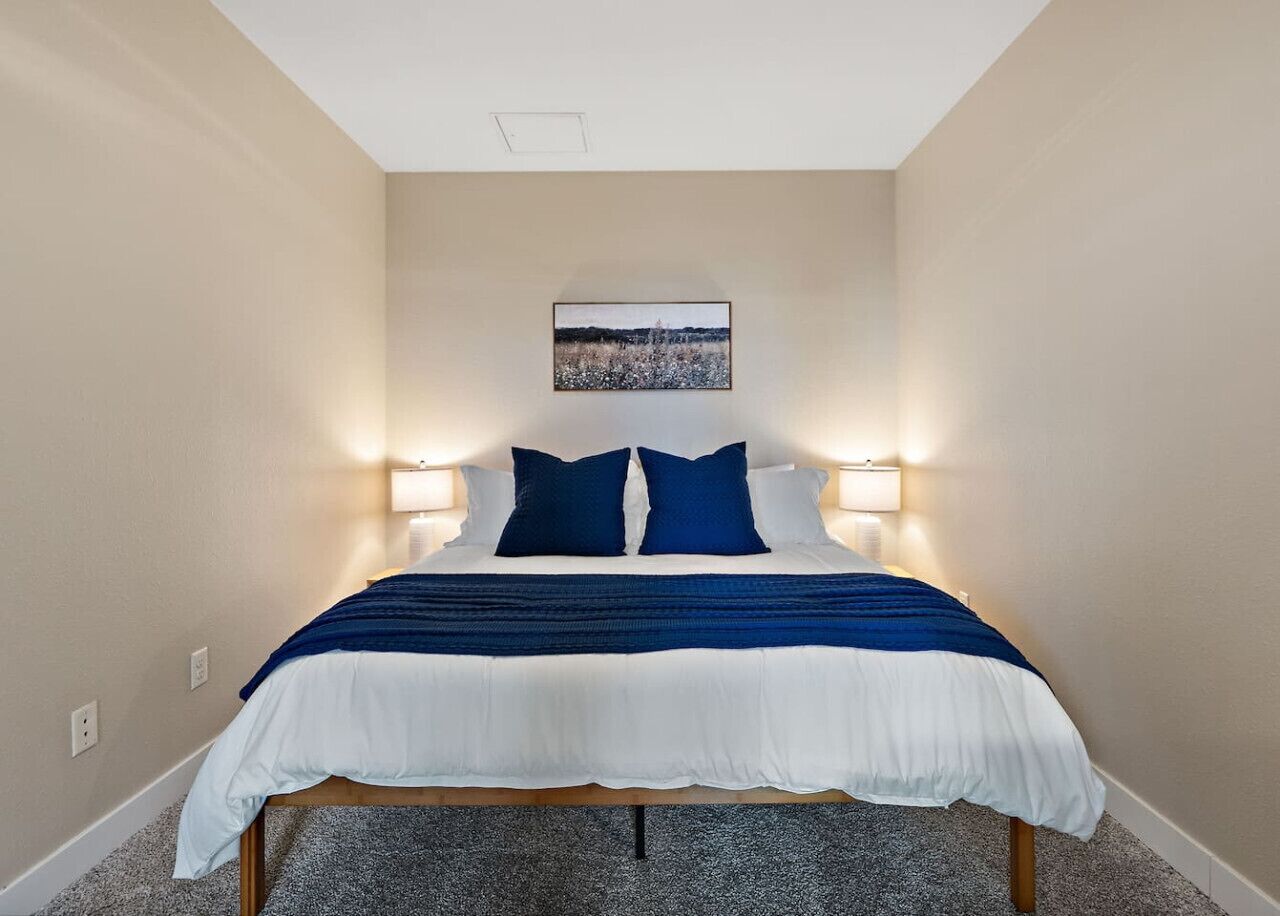 A bedroom with a bed, two bedside lamps, a framed picture on the wall, and a carpeted floor.