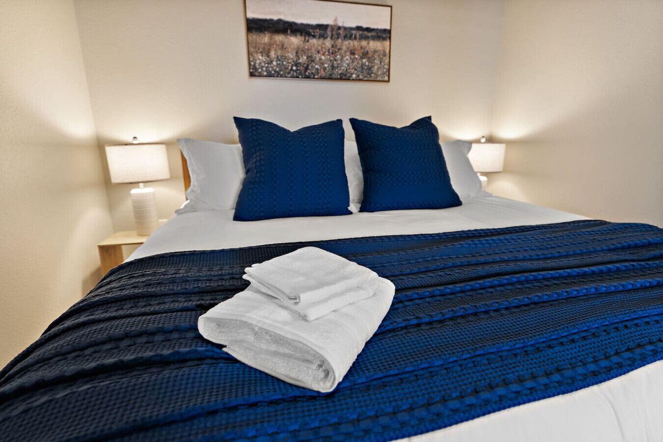 Una cama bien hecha con ropa de cama azul y blanca, una toalla blanca doblada y un cuadro de paisaje en la pared.