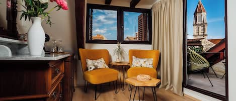 A room with a wooden floor, a white fluffy rug, two yellow chairs with floral cushions, a white vase with flowers, and a view of a building with a tower through a large window.