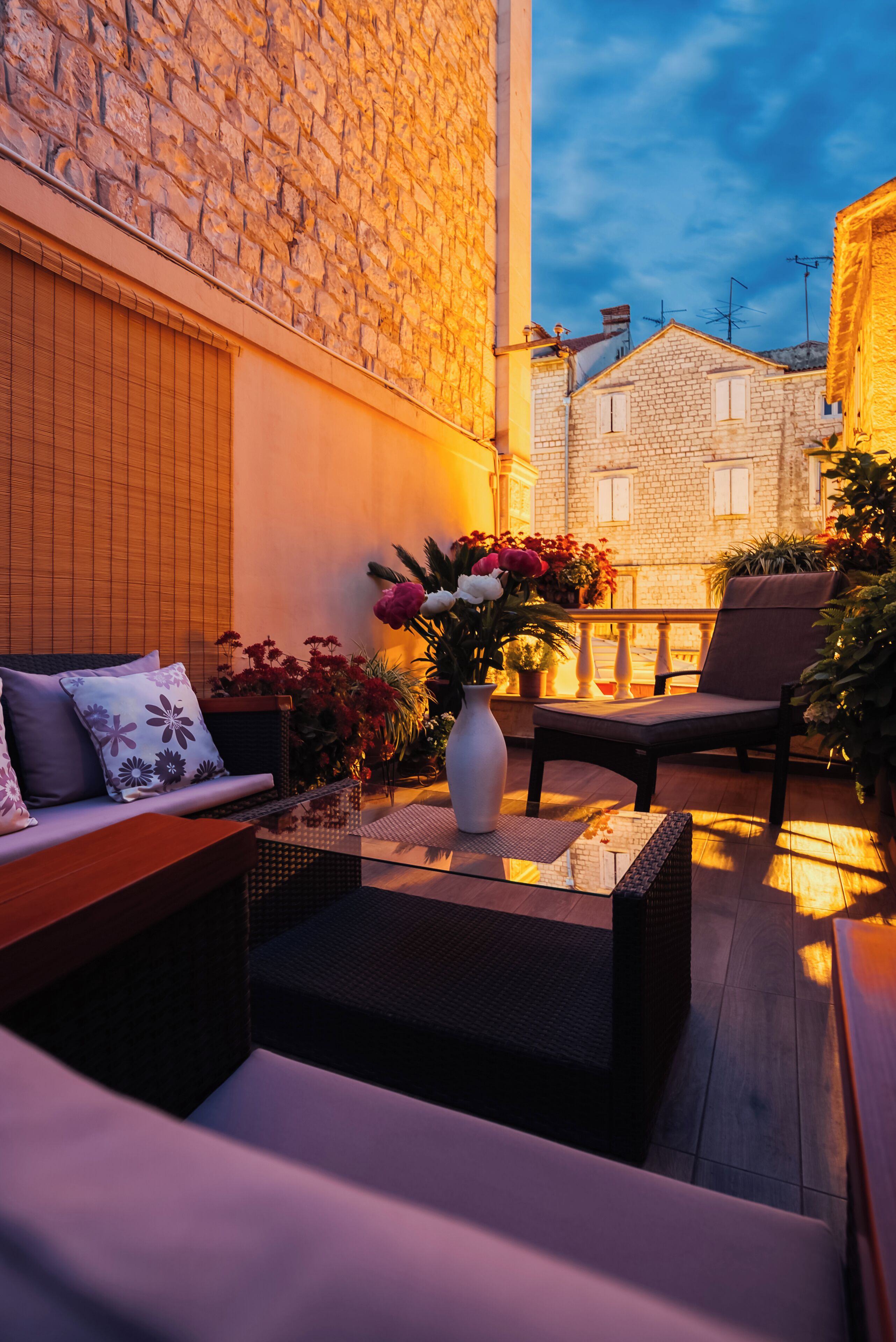 A cozy outdoor seating area with a sofa, chairs, and a table, surrounded by potted plants and a vase of flowers.
