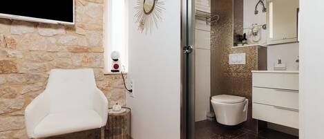 A bathroom with a shower, toilet, and sink. There is a white chair and a small table with a decorative item.