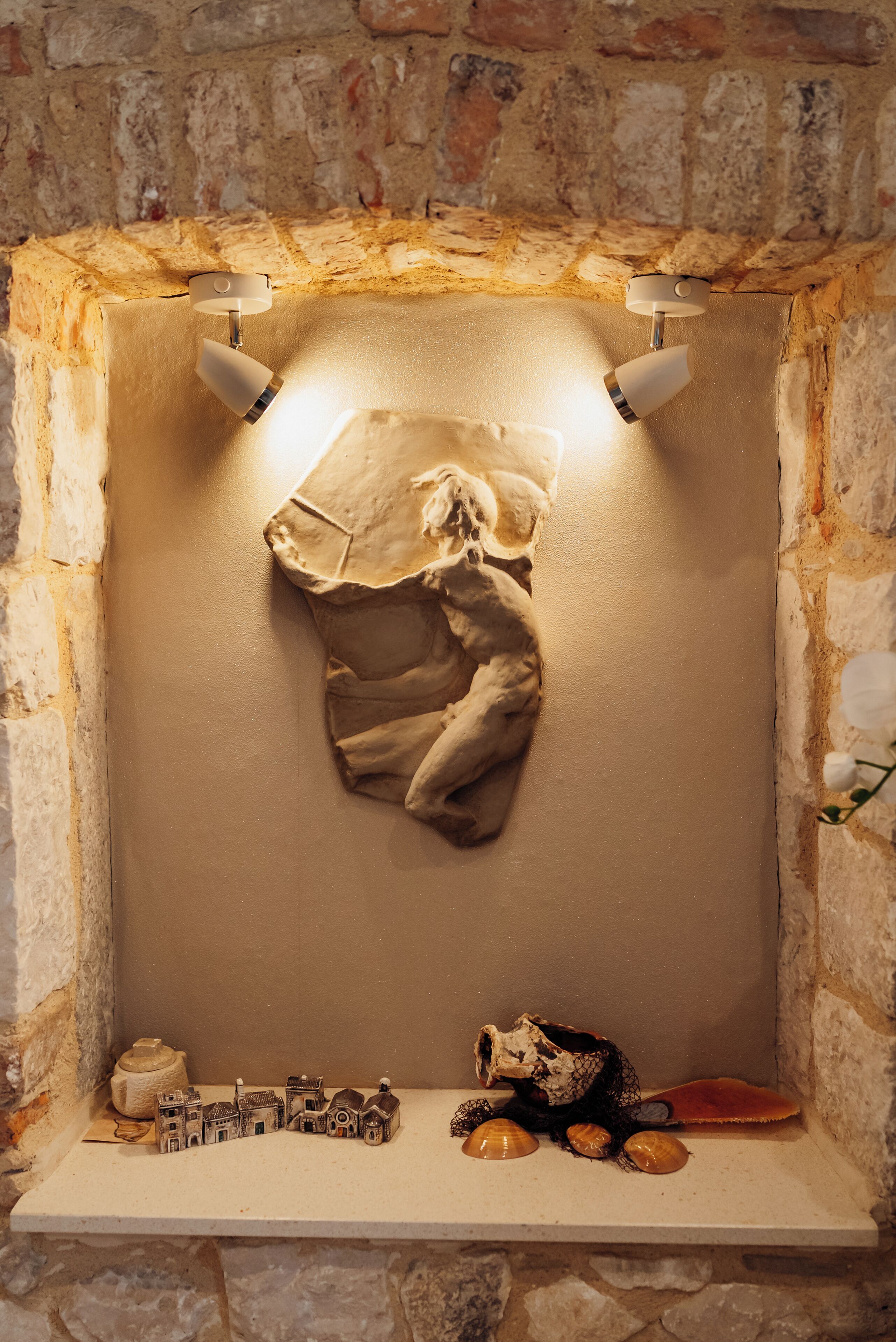 Um recanto com paredes de pedra, um tecido pendurado, uma prateleira com objetos decorativos e duas luzes fixadas na parede.
