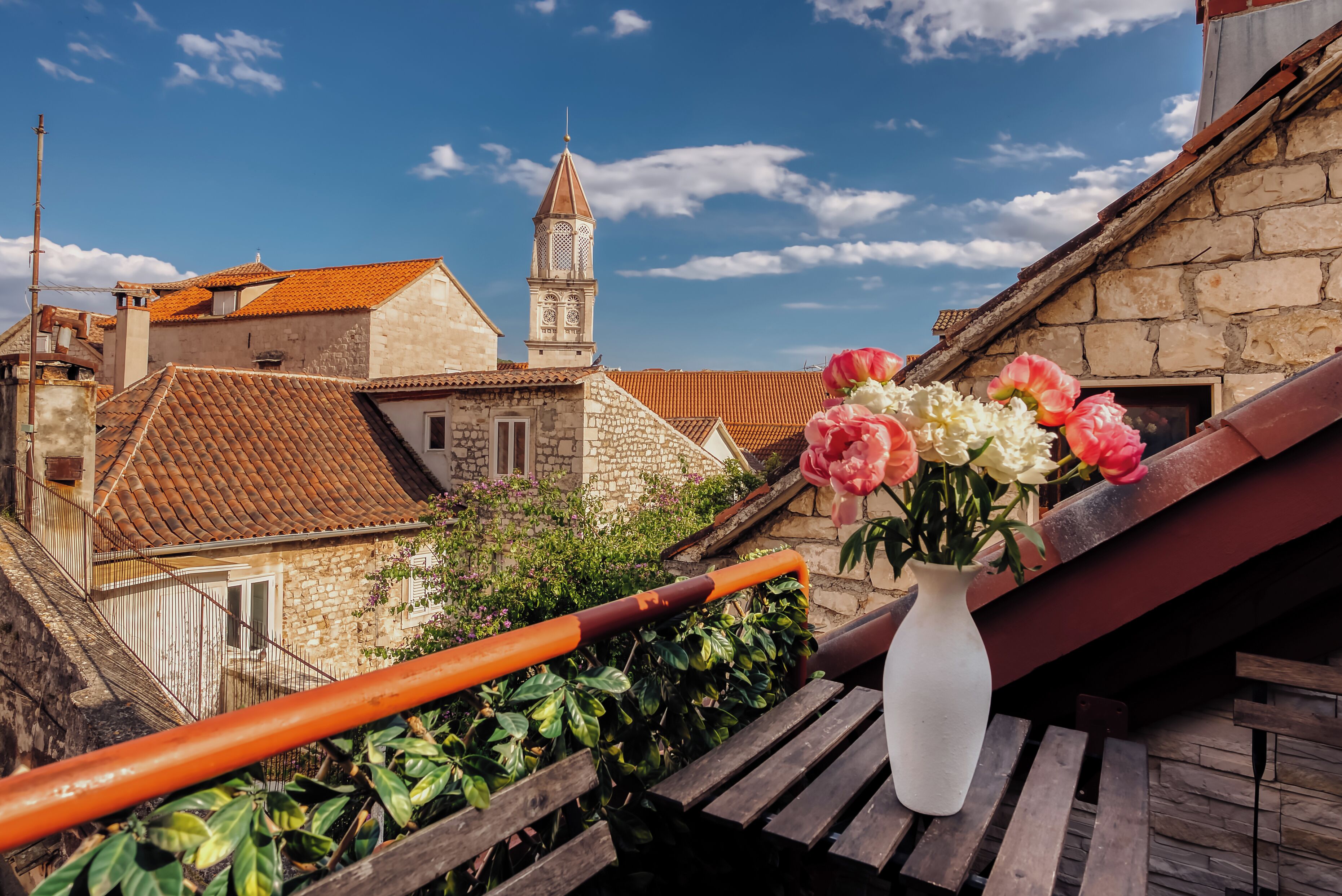 Un balcon orné d’un vase blanc rempli de fleurs, doté d’une balustrade en bois, et offrant une vue sur un bâtiment en pierre surmonté d’une tour.