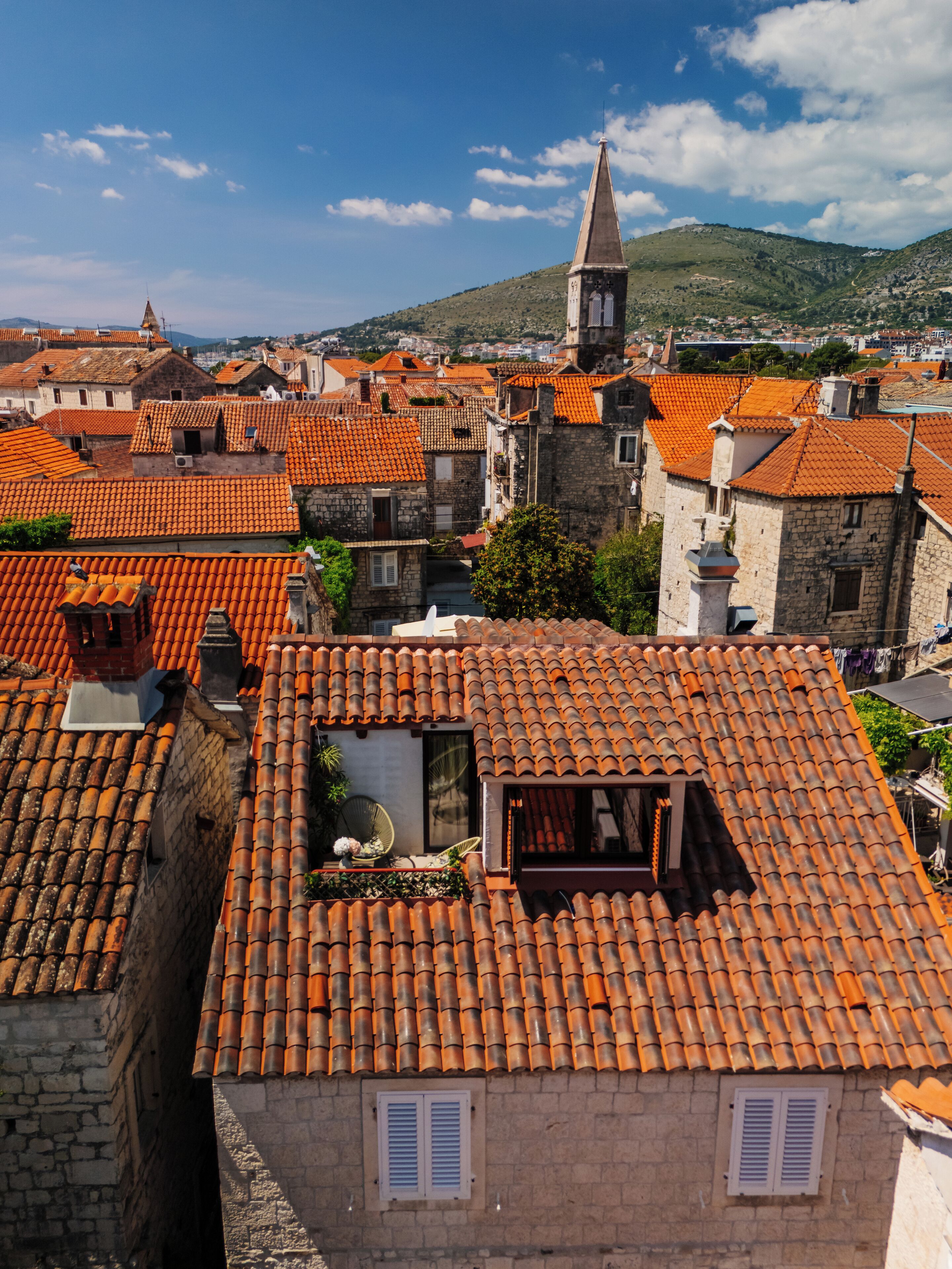 A view of a town with red-tiled roofs and stone buildings, including a church with a tall steeple.